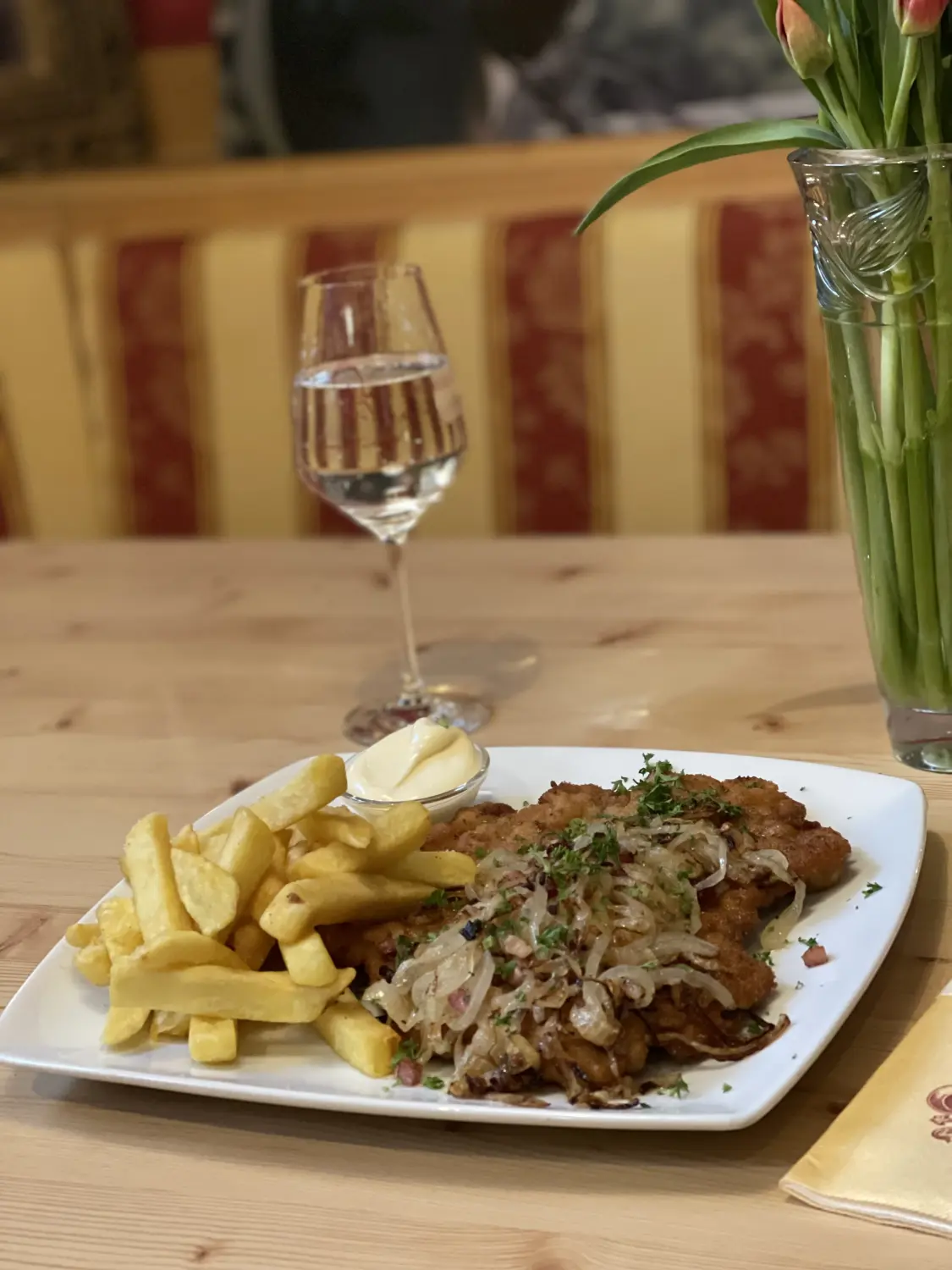 Schnitzel mit Pommes frites und sautierten Zwiebeln, eventuell mit einer Sauce wie Mayonnaise oder Aioli und einem Glas Wasser. Restaurant Topinambur