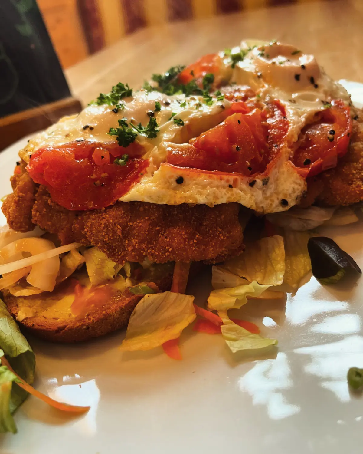 Eine Variation eines Hähnchen- oder Schweineschnitzelsandwiches, wahlweise offen serviert, mit Spiegeleiern und Tomatenscheiben, garniert mit Petersilie und schwarzem Sesam vom Restaurant Topinambur